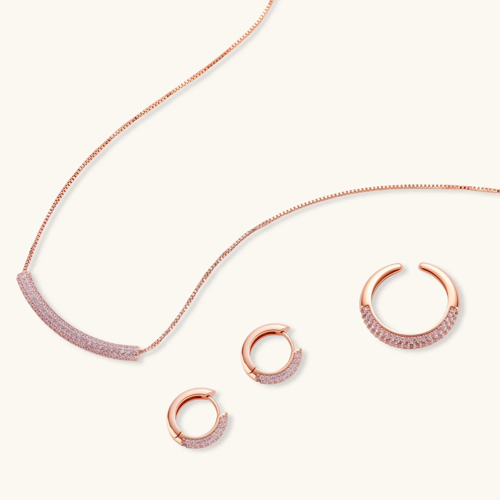 Rose gold jewelry set with pavé cubic zirconia stones including curved necklace, hoop earrings, and adjustable ring on cream background.