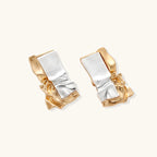 Close-up of avant-garde sculptural earrings with flowing liquid metal design in gold and silver tones on white background.
