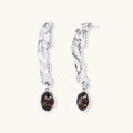 Silver-toned twisted metal earrings with dark brown oval resin stones dangling from sculptural posts on white background.