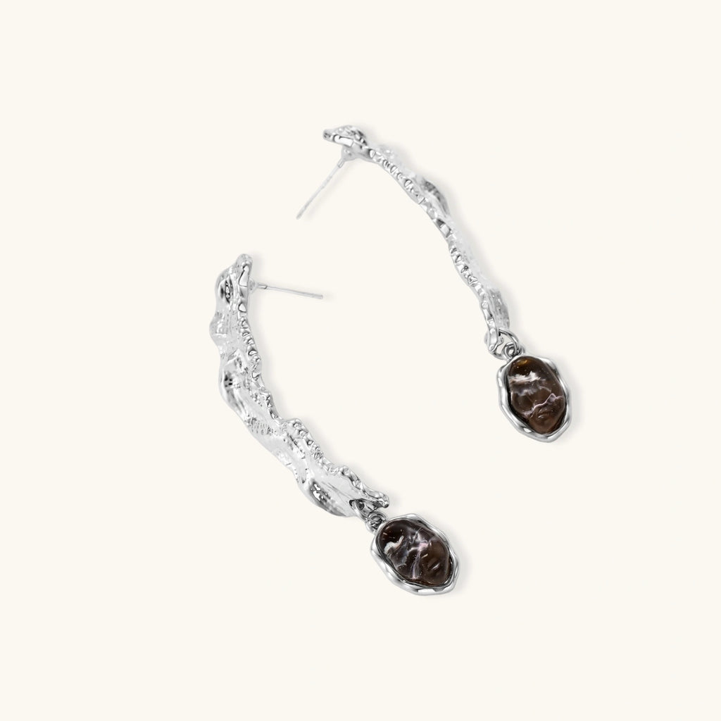 Silver-toned twisted metal earrings with dark brown oval resin stones dangling from sculptural posts on white background.
