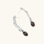 Silver-toned twisted metal earrings with dark brown oval resin stones dangling from sculptural posts on white background.