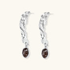 Silver-toned twisted metal earrings with dark brown oval resin stones dangling from sculptural posts on white background.