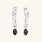 Silver-toned twisted metal earrings with dark brown oval resin stones dangling from sculptural posts on white background.