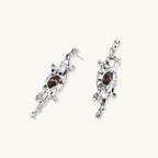 Flowing silver-toned earrings with organic liquid shapes and embedded brown resin stones on white background.