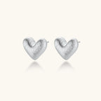 Pair of brushed platinum-plated 925 sterling silver puffy heart stud earrings on a cream background.